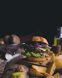 A gourmet burger loaded with layers of fresh lettuce, red onion, tomato, pickle slices, cheese, and a beef patty sits on a wooden board. Rustic potato wedges are scattered around, and a small glass bottle of olive oil along with a sauce container can be seen in the background. A metal bowl with raw potatoes and a small cup with seasoning are also visible, contributing to a rustic, cozy kitchen atmosphere.
