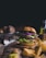A gourmet burger loaded with layers of fresh lettuce, red onion, tomato, pickle slices, cheese, and a beef patty sits on a wooden board. Rustic potato wedges are scattered around, and a small glass bottle of olive oil along with a sauce container can be seen in the background. A metal bowl with raw potatoes and a small cup with seasoning are also visible, contributing to a rustic, cozy kitchen atmosphere.