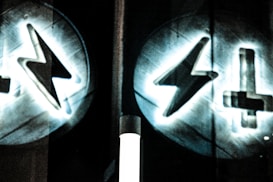 Two circular symbols featuring a lightning bolt and an upside-down cross are projected onto a dark surface. The symbols are surrounded by a glowing halo effect, creating a mysterious and slightly ominous atmosphere.