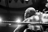 Two athletes, covered in sweat, are engaged in a close contact moment in a boxing ring. The image is in black and white, with intense overhead lighting creating strong contrasts and highlighting the athletes' muscular physiques.