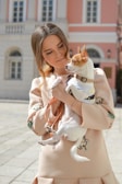 A young woman is holding a small dog, possibly a Chihuahua, in her arms. She has long hair and is wearing a beige outfit with embroidery. The dog is wearing a harness and leash. The background features a pink and white building with large windows.