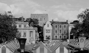 A historic cityscape featuring a castle with a flag on top is visible in the background, surrounded by trees. The foreground showcases a collection of market stalls with striped rooftops. Elegant brick and stone buildings with traditional architecture line the middle ground, displaying a Lloyds Bank and other commercial signage.