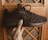 A hand holds a black athletic shoe against a wooden background with a lattice pattern. The shoe has a sleek design with mesh fabric and visible stitching.