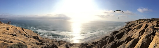 A breathtaking coastal view with rugged cliffs overlooking a vast ocean. The sunlight is reflecting off the water, creating a bright, shimmering effect. A paraglider is soaring in the sky, adding an adventurous element to the serene landscape.