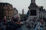 A crowded city square with people walking and gathering around a large monument. Iconic architecture can be seen in the background, including a clock tower. The square is surrounded by historic buildings, and there's a red double-decker bus in the scene.