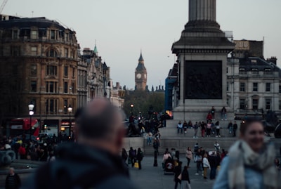 A crowded city square with people walking and gathering around a large monument. Iconic architecture can be seen in the background, including a clock tower. The square is surrounded by historic buildings, and there's a red double-decker bus in the scene.