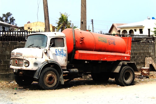 An old, weathered truck with a white cab and a large red tank attached to its back. The truck is parked on a dirt road beside a tall concrete wall. It has visible rust and graffiti on the side. The background includes utility poles, buildings with various roof styles, and some greenery.