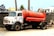 An old, weathered truck with a white cab and a large red tank attached to its back. The truck is parked on a dirt road beside a tall concrete wall. It has visible rust and graffiti on the side. The background includes utility poles, buildings with various roof styles, and some greenery.