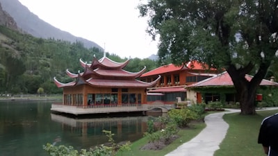 A scenic lakeside setting with a traditional-style wooden building with an ornate, multi-tiered roof. The structure is surrounded by lush greenery, including trees and a groomed lawn, with a mountain range in the background. A winding path leads to the building.