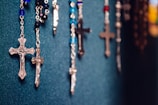 Several rosaries with crucifixes are hanging against a textured dark teal background. The rosaries feature beads in a variety of colors, including blue, black, and red. The crucifixes are metallic, varied in size, and show intricate detailing.