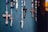 Several rosaries with crucifixes are hanging against a textured dark teal background. The rosaries feature beads in a variety of colors, including blue, black, and red. The crucifixes are metallic, varied in size, and show intricate detailing.