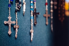 Several rosaries with crucifixes are hanging against a textured dark teal background. The rosaries feature beads in a variety of colors, including blue, black, and red. The crucifixes are metallic, varied in size, and show intricate detailing.