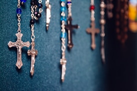 Several rosaries with crucifixes are hanging against a textured dark teal background. The rosaries feature beads in a variety of colors, including blue, black, and red. The crucifixes are metallic, varied in size, and show intricate detailing.