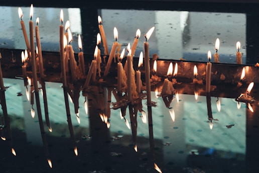 A series of lit, wax candles standing upright and melted down along a reflective surface that resembles water. The candles' flickering flames emit a warm glow, while their reflections create a serene mirror effect.