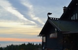 A dark wooden house with ornate architectural details sits on the right side against a backdrop of a vast, scenic landscape. The sky is filled with a mix of soft clouds and warm hues of sunset or sunrise, casting a serene light over the silhouette of a forested horizon.