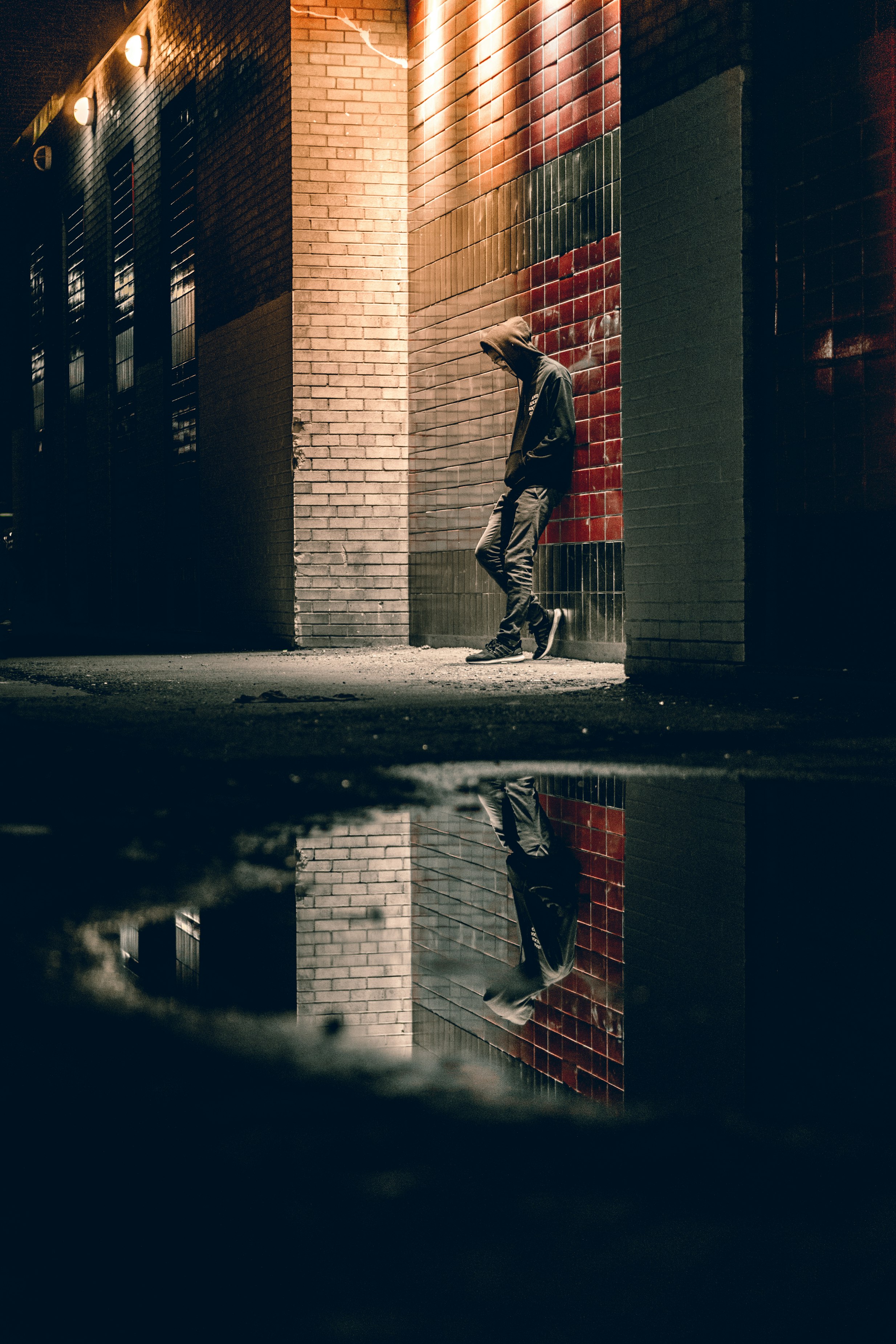 A photograph of a person leaning against a brick wall with their face covered by a hooded sweatshirt. In the foreground is a puddle that reflects the person.