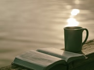 A close-up of a wooden deck with a cup of coffee and an open book beside the water.