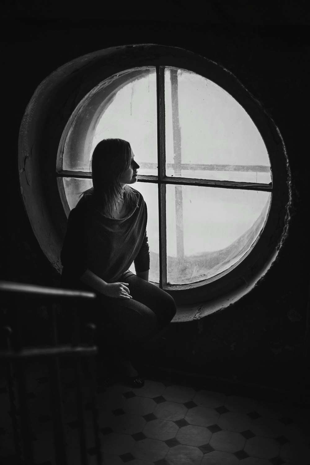 grayscale photo of woman sits near round wooden framed window