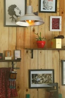 A cozy and eclectic interior with wooden wall panels adorned by various framed artworks and photographs. A hanging industrial-style lamp with a visible bulb provides warm lighting. Shelves hold a red potted plant and small decorative items. A colorful hanging fabric with tassels adds a touch of vibrancy.