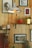 A cozy and eclectic interior with wooden wall panels adorned by various framed artworks and photographs. A hanging industrial-style lamp with a visible bulb provides warm lighting. Shelves hold a red potted plant and small decorative items. A colorful hanging fabric with tassels adds a touch of vibrancy.