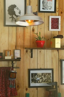 A cozy and eclectic interior with wooden wall panels adorned by various framed artworks and photographs. A hanging industrial-style lamp with a visible bulb provides warm lighting. Shelves hold a red potted plant and small decorative items. A colorful hanging fabric with tassels adds a touch of vibrancy.