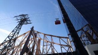 A red cable car travels along suspended cables next to a steel bridge structure, which appears to be part of a larger bridge featuring brownish metalwork against a clear blue sky. In the foreground, the glass facade of a tall building reflects part of the bridge and the sky.
