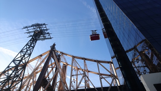 A red cable car travels along suspended cables next to a steel bridge structure, which appears to be part of a larger bridge featuring brownish metalwork against a clear blue sky. In the foreground, the glass facade of a tall building reflects part of the bridge and the sky.