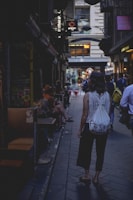 A narrow alleyway lined with small shops and cafes. People are casually walking or sitting outside, creating a bustling yet cozy atmosphere. The lighting is subdued, contributing to a moody, urban vibe.
