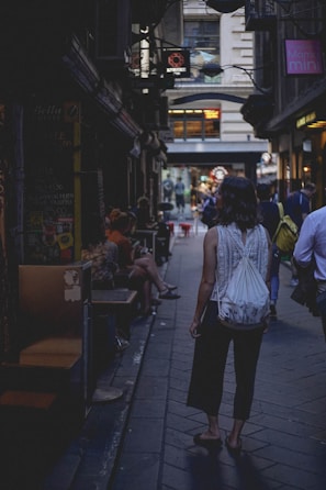 A narrow alleyway lined with small shops and cafes. People are casually walking or sitting outside, creating a bustling yet cozy atmosphere. The lighting is subdued, contributing to a moody, urban vibe.