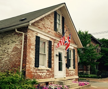 A small, charming brick house with black shutters and a white door. Two flags, including the American and British flags, are prominently displayed at the front. Vibrant pink and purple flowers line the front yard, and the background features a tree and a neighboring building.