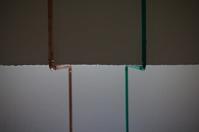 Two vertical pipes, one copper-colored and the other teal, are on a background that is divided horizontally, with dark gray on top and light gray below.