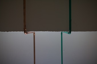 Two vertical pipes, one copper-colored and the other teal, are on a background that is divided horizontally, with dark gray on top and light gray below.