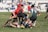 Players in a rugby match are engaged in a tackle on a grassy field, with athletes wearing jerseys in red and green. Spectators can be seen in the background on bleachers.