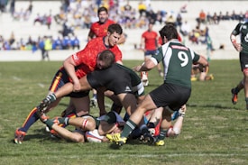 Players in a rugby match are engaged in a tackle on a grassy field, with athletes wearing jerseys in red and green. Spectators can be seen in the background on bleachers.