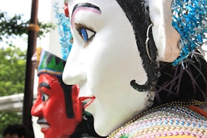 Several large, colorful effigies or puppets with detailed facial features are pictured. The effigy in the foreground has a prominently white face with detailed black, red, and blue facial markings and decorations. The effigy behind has a striking red face with black and white decorations. Both appear to be part of a larger display or parade, with trees and other items slightly visible in the background.