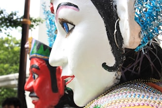 Several large, colorful effigies or puppets with detailed facial features are pictured. The effigy in the foreground has a prominently white face with detailed black, red, and blue facial markings and decorations. The effigy behind has a striking red face with black and white decorations. Both appear to be part of a larger display or parade, with trees and other items slightly visible in the background.