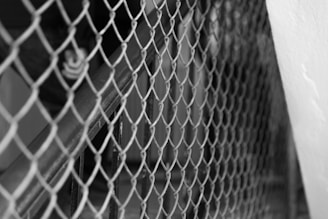 A close-up view of a metal chain-link fence. The fence is constructed of interlocking metal wires forming a diamond pattern. Behind the fence, there is a handrail and some blurred objects, indicating depth and possibly an underlying staircase or passage.