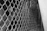 A close-up view of a metal chain-link fence. The fence is constructed of interlocking metal wires forming a diamond pattern. Behind the fence, there is a handrail and some blurred objects, indicating depth and possibly an underlying staircase or passage.