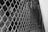 A close-up view of a metal chain-link fence. The fence is constructed of interlocking metal wires forming a diamond pattern. Behind the fence, there is a handrail and some blurred objects, indicating depth and possibly an underlying staircase or passage.