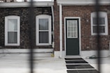 A brick wall facade featuring two windows and a central door with a glass panel. The windows have white frames and blinds. A black hose and a few black mats are visible on the white ground in front of the facade. There is a pipe running vertically next to the door, and a small outdoor lamp is mounted on the wall above the door.