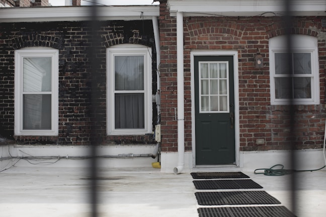 A brick wall facade featuring two windows and a central door with a glass panel. The windows have white frames and blinds. A black hose and a few black mats are visible on the white ground in front of the facade. There is a pipe running vertically next to the door, and a small outdoor lamp is mounted on the wall above the door.