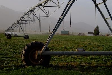 A large irrigation system with metal structures and wheels spans across a green field, with a backdrop of small buildings and distant mountains under a misty sky.