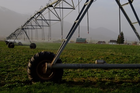 A large irrigation system with metal structures and wheels spans across a green field, with a backdrop of small buildings and distant mountains under a misty sky.