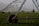 A large irrigation system with metal structures and wheels spans across a green field, with a backdrop of small buildings and distant mountains under a misty sky.