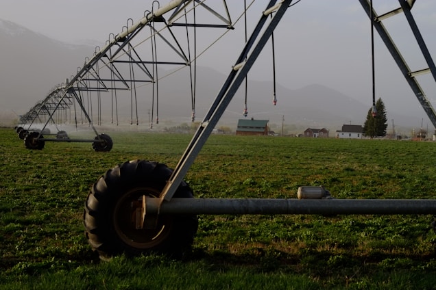 A large irrigation system with metal structures and wheels spans across a green field, with a backdrop of small buildings and distant mountains under a misty sky.