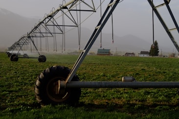 A large irrigation system with metal structures and wheels spans across a green field, with a backdrop of small buildings and distant mountains under a misty sky.