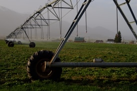 A large irrigation system with metal structures and wheels spans across a green field, with a backdrop of small buildings and distant mountains under a misty sky.