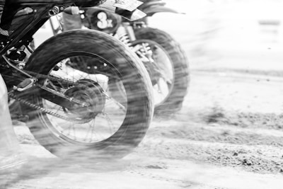 Two motorcycles are captured in motion with the focus on their rear wheels, kicking up dirt and debris. The image is in black and white, emphasizing the motion blur and texture of the terrain.