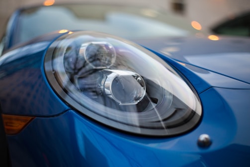 A close-up view of a car's headlight, showcasing its sleek design and reflective surface. The headlight is part of a blue vehicle with a modern and shiny finish. The background is blurred, hinting at an urban setting with faint lights and structural outlines.