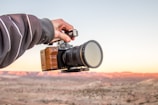 A hand is holding a professional camera with a wooden grip against a backdrop of a desert landscape at sunset. The sky is clear with subtle hues of orange and pink reflecting on the terrain.